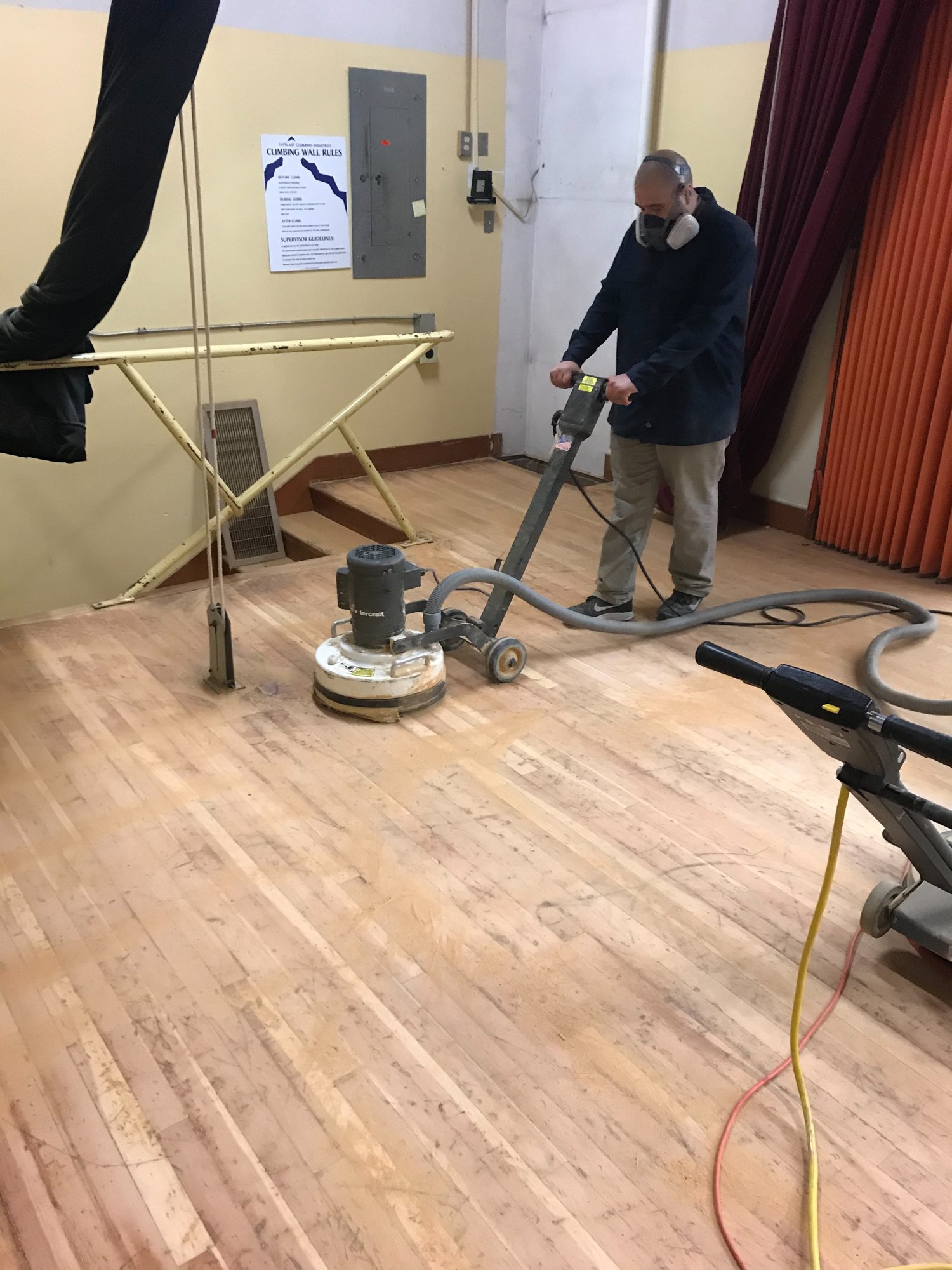 TBM crew sanding a wood gym floor with a floor grinder, wearing a respirator
