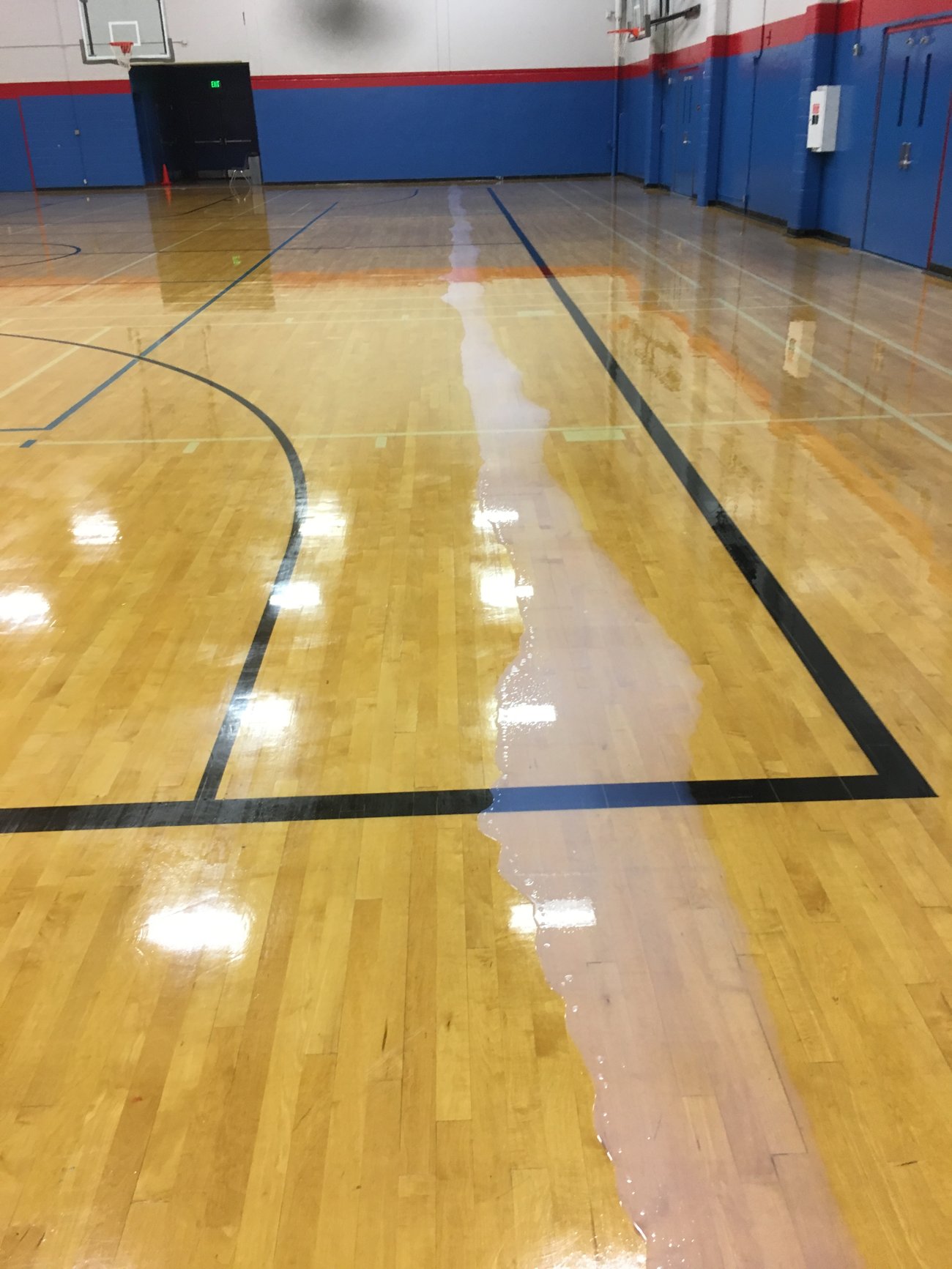 Gym floor mid-refinish — the wet finish line splits the dull worn floor from the fresh mirror gloss